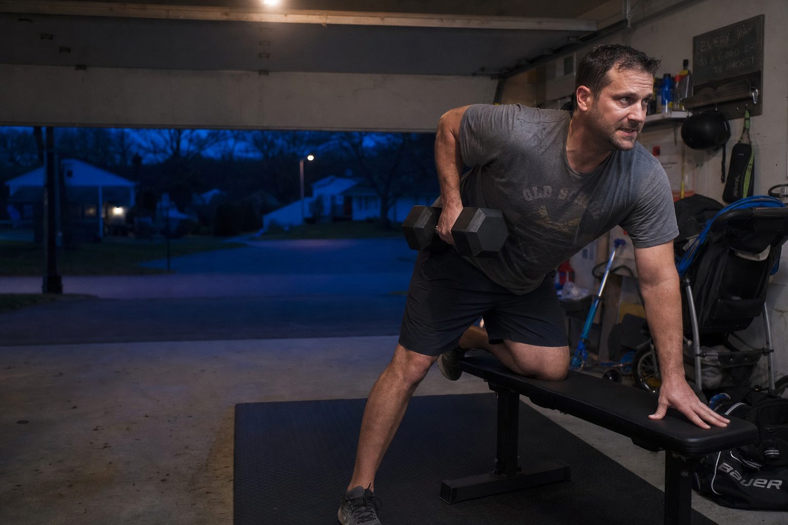 A dad training with a dumbbell in his garage before dawn, garage door open to a dark street